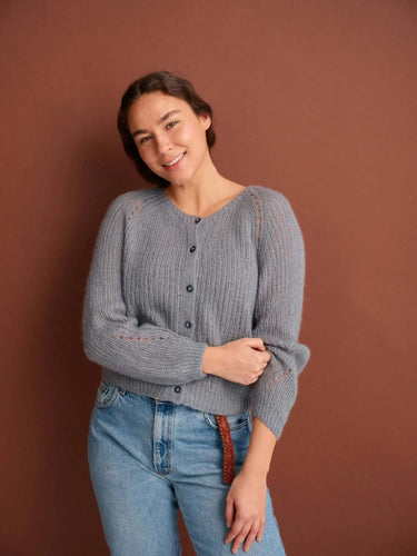 Model wearing a grey knitted sweater from 52 Weeks of Sweaters book, available for pre-order, smiling against brown background