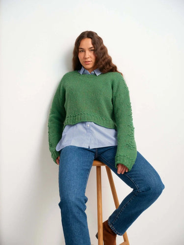 Woman wearing green sweater from "52 Weeks of Sweaters" knitting book collection, seated on stool.