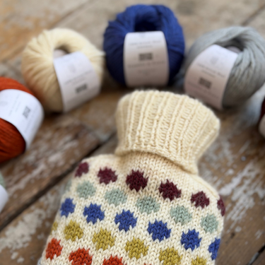 A cream-colored knitted hot water bottle cover with a colorful pattern of dots, displayed on a rustic wooden surface with skeins of yarn in the background.