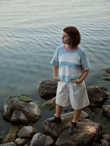 Woman standing on rocks by water, wearing a stylish blue and white sweater featured in Laine Magazine Spring 28 issue.