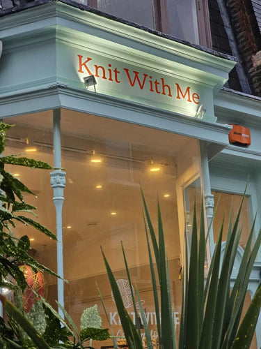 Storefront with 'Knit With Me' sign above glass door, surrounded by plants.