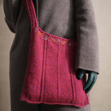 Person wearing a grey coat and green gloves holding a pink knit bag with beads, showcasing Beaded Colourwork knitting technique.