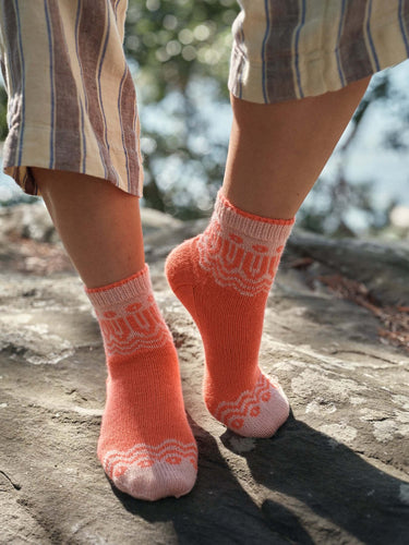 Person wearing orange knit socks from Laine Magazine Spring Issue 28 collection, standing on sunlit rocky beach.