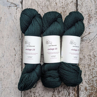 Three skeins of Wool Decanted Vintage 23 4ply Yarn in dark green on a wooden surface.