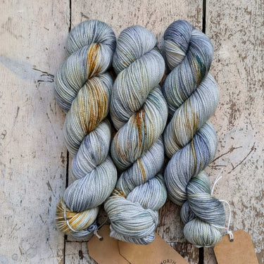 Three skeins of A&P Plump Sock DK non-mulesed merino yarn hand dyed in grey and gold for soft, durable knit socks and accessories.