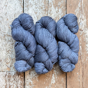 Three skeins of Manos Maxima extrafine merino wool yarn in dark gray on rustic wood background.
