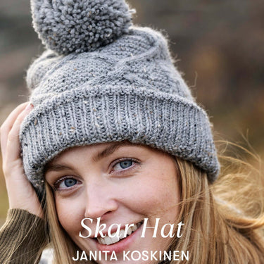 Woman wearing Skar Hat, grey knit hat with pom-pom, by Janita Koskinen, smiling in outdoors setting.
