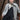 Woman wearing a gray cardigan over a black outfit with a patterned scarf, standing against a wooden wall. Tukubook knitting book