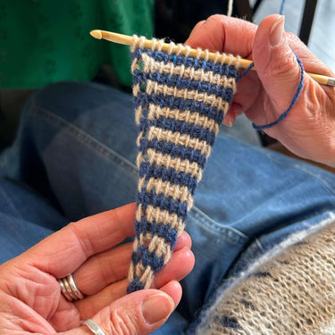 Person knitting a small striped fabric with blue and beige yarn.