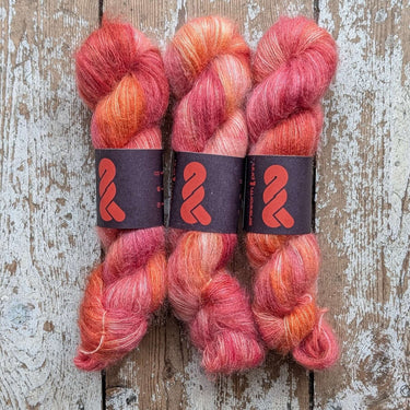 Three skeins of hand dyed A&P Lace Silk Mohair in shades of pink and orange on a rustic wood surface.