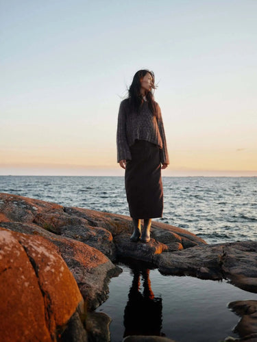 Woman wearing Art of the Sea knitwear standing on rocks by the sea during sunset.