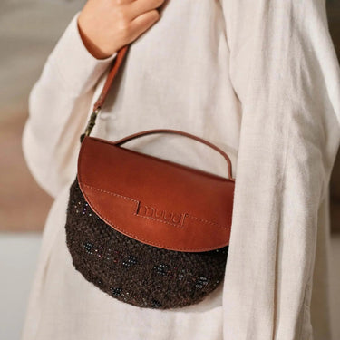 Brown leather half-moon handbag with beaded detailing held by model in beige shirt.