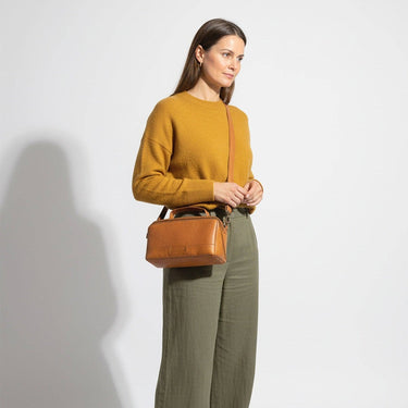 Woman holding a brown handbag wearing a mustard yellow sweater and green pants on a white background