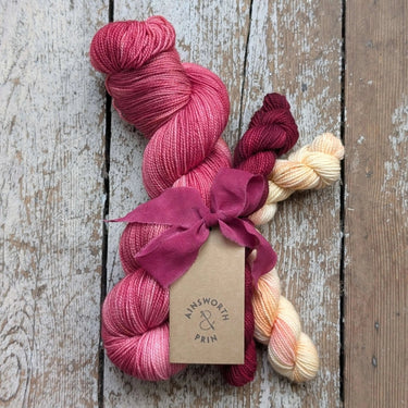 Ainsworth & Prin Flower Yarn Knitting Kits with pink skeins and silk ribbon on a wooden background.