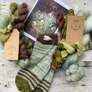Flower Yarn Knitting Kits by Ainsworth & Prin with skeins, silk ribbon, and sock detail in earthy tones placed on a rustic background.