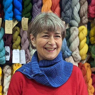 Woman in a red sweater wearing a blue knitted scarf in front of colorful yarns, promoting Beginner Knitting Class.