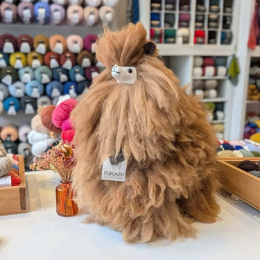 Inkari Stuffed Alpaca Natural size displayed on a shop counter with colorful yarn background