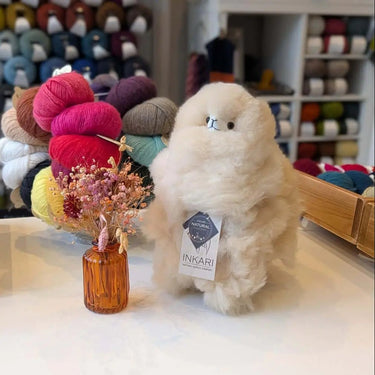 Stuffed Alpaca toy from Inkari displayed on a table with colorful yarn and a decorative vase, showcasing the Natural type.
