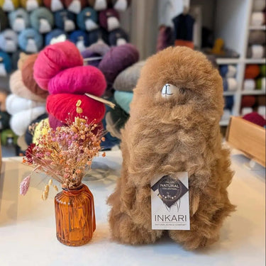 Inkari Stuffed Alpaca Animal in brown, displayed with colorful wool in background and decorative vase