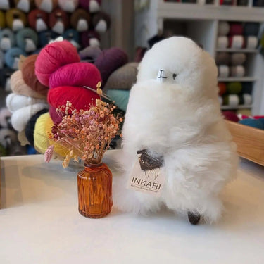 Stuffed Alpaca by Inkari next to a vase, soft and fluffy, perfect for cuddling. Available in various sizes and colors.