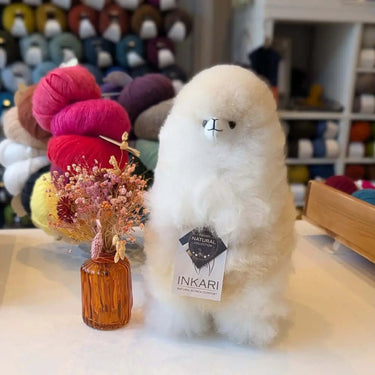 Natural Inkari Stuffed Alpaca in a cozy yarn shop setting, showcasing its soft fur and plush design alongside a decorative vase of flowers.