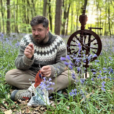 Instructor in a forest setting teaching Yarn Spinning class with a spinning wheel, surrounded by purple flowers.
