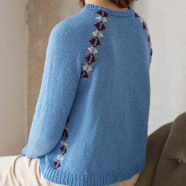 Woman wearing a blue knitted sweater with floral patterns on the back, sitting against a neutral wall.