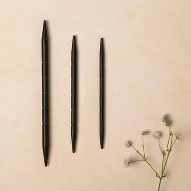 Set of three ebony wooden Lantern Moon cable needles in different sizes on a beige surface with decorative flowers.