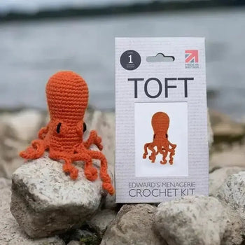 Orange crochet octopus kit for "Crochet your first Amigurumi Workshops" against a rocky background near water.