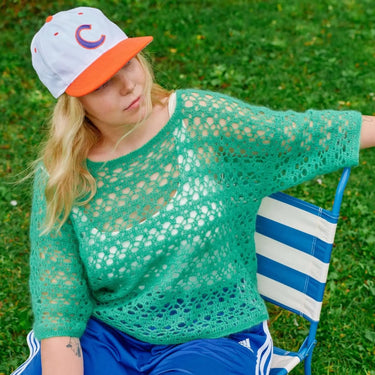 Person wearing a crocheted green top from Molla Mills' Hooks Out collection, sitting in a striped chair outdoors.