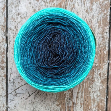 Gradient hand-dyed Snek merino wool yarn with blue-green hues on wooden background.