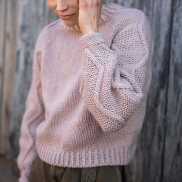 Woman wearing a light pink knitted sweater against a wooden background. Tukubook knitting book