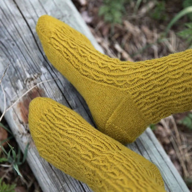 Mustard yellow knitted socks on a wooden log with greenery in the background. Tukubook knitting book