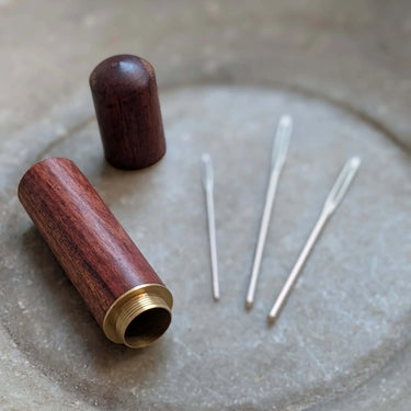 Wooden darning needle case with 3 metal darning needles on table.