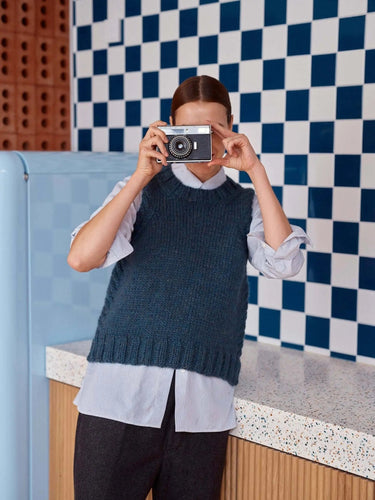 Person wearing a knitted sweater holding a camera, related to 52 Weeks of Sweaters pre-order knitting book publication.
