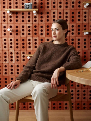 Woman wearing a brown sweater from the "52 Weeks of Sweaters" knitting book collection, seating against a warm-toned textured wall.