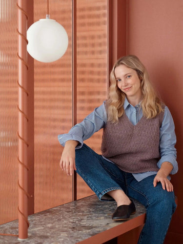 Woman sitting in a modern room wearing a knit vest, showcasing style from "52 Weeks of Sweaters" knitting book available for pre-order.