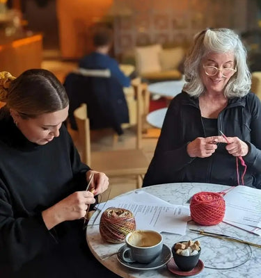 Two women sitting at a table in a cozy café, both knitting 