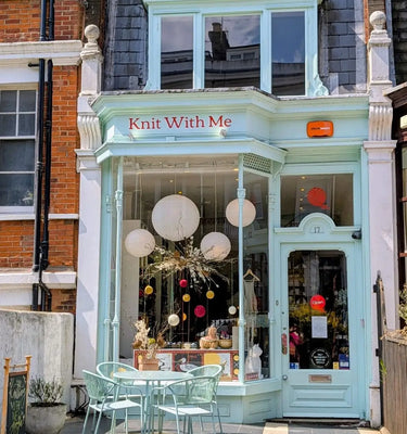 Light blue storefront with 'Knit With Me' sign, glass windows, and outdoor seating area.