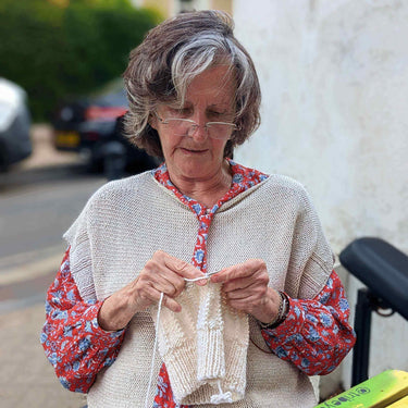 Woman knitting outdoors, part of a Beginner Knitting Class, wearing glasses and a colorful outfit, focused on her knitting project.