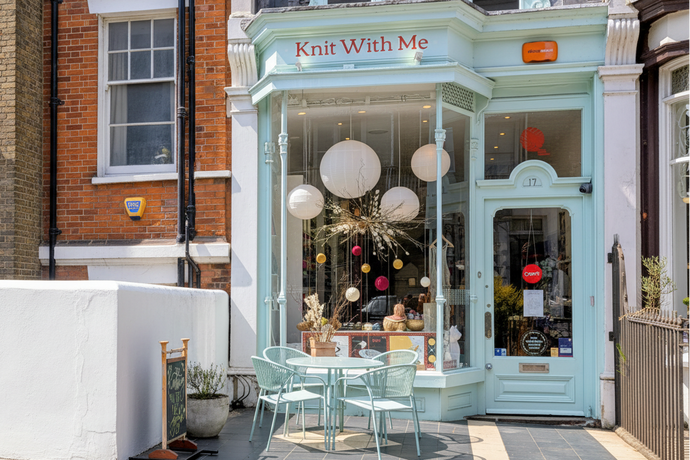 Storefront with 'Knit With Me' sign, outdoor seating area, and decorative elements.