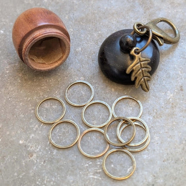Wooden acorn stitch marker case with 10 ring stitch markers and clasp leaf charm on a grey surface.