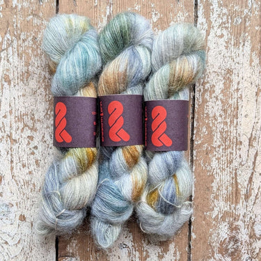 Three skeins of A&P Lace Silk Mohair yarn, hand dyed in soft hues, displayed on a rustic wooden surface.