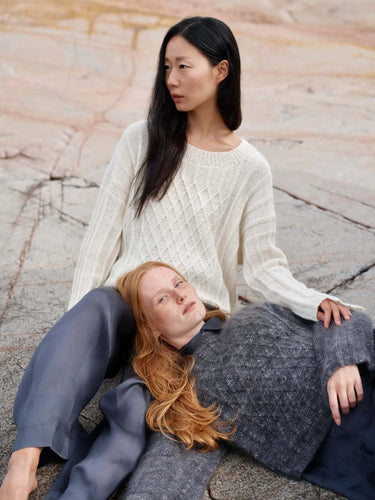 Two models wearing knitwear from Art of the Sea collection, showcasing intricate Moomins-inspired designs, sitting on rocky terrain.