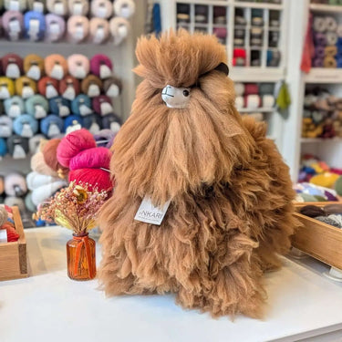 Inkari Stuffed Alpaca Animal in Monsterfluff style, brown color, displayed on a shop counter with yarn background.