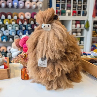 Stuffed Alpaca animal by Inkari on display, showcasing its fluffy brown coat, surrounded by colorful yarn skeins.