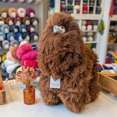 Natural Inkari Stuffed Alpaca Animal in a store display, showcasing its fluffy brown fur and soft texture amid colorful yarns.