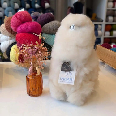 Inkari Stuffed Alpaca next to a vase with dried flowers, surrounded by colorful yarn balls in a cozy shop setting