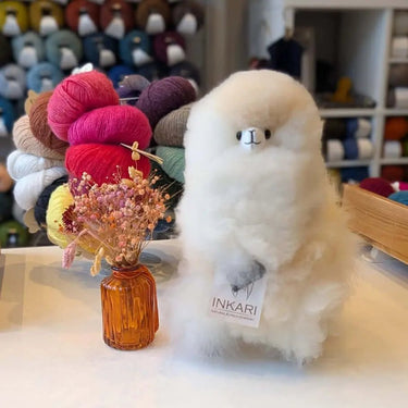 Inkari Stuffed Alpaca Animal in white, fluffy texture, displayed on a table next to a vase of flowers.
