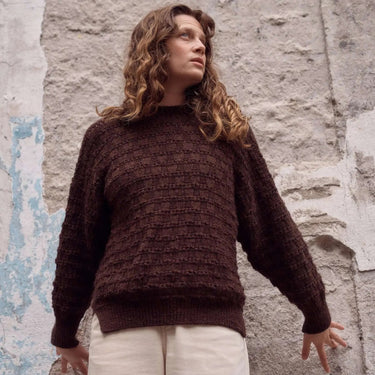 Woman wearing a brown knitted sweater and white pants against a textured stone wall.